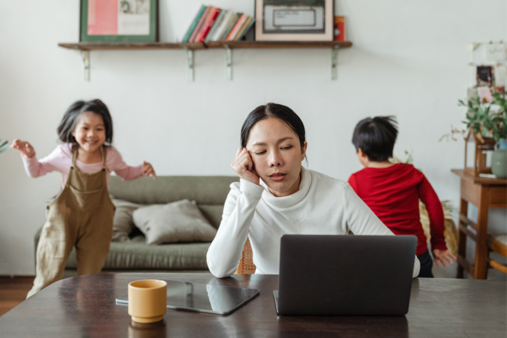 Tired bilingual mother working on computer while kids are running around behind her, wondering if their family should drop a language